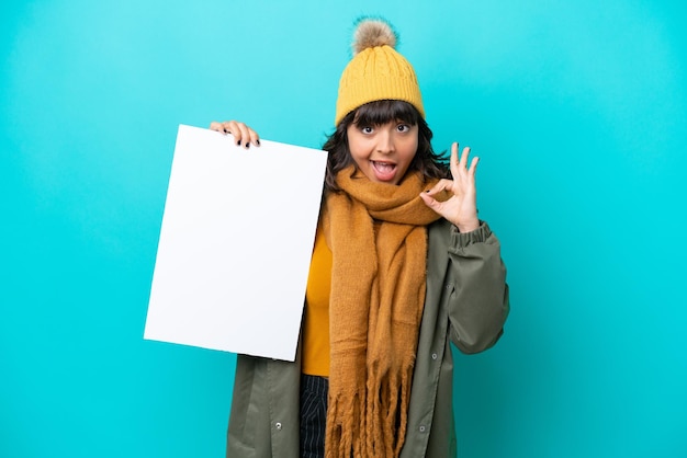 Молодая латинская женщина в зимней куртке на синем фоне держит пустой плакат и делает знак ОК