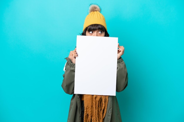 Young latin woman wearing winter jacket isolated on blue background holding an empty placard and hiding behind it