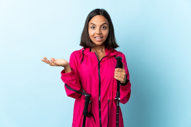 Young latin woman with backpack and trekking poles isolated on blue background with shocked facial expression