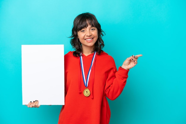 Young latin woman with medals isolated on blue background holding an empty placard and pointing side