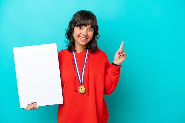 Young latin woman with medals isolated on blue background holding an empty placard with surprised expression