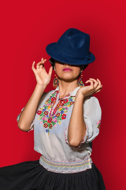 Young latin woman with traditional dress covering part of her face with a blue hat in red background