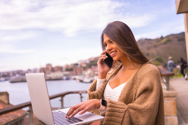 A young Latin woman with a white tshirt and a brown wool sweater teleworking on the terrace of a house by the sea smiling and enjoying the working day