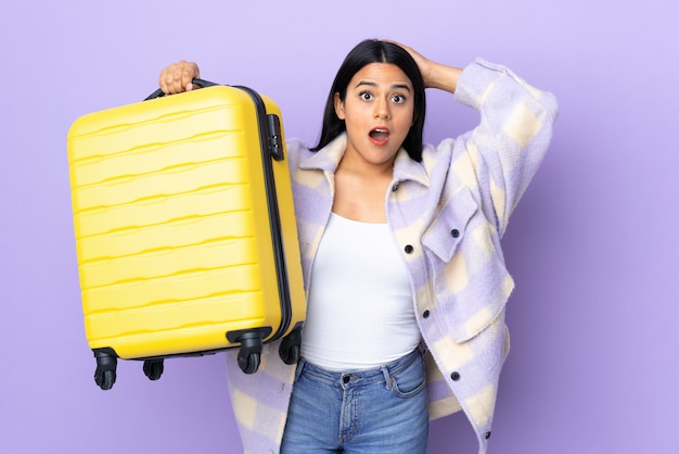 Young latin woman woman isolated on purple in vacation with travel suitcase and surprised