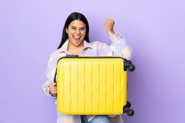 Young latin woman woman isolated on purple wall in vacation with travel suitcase