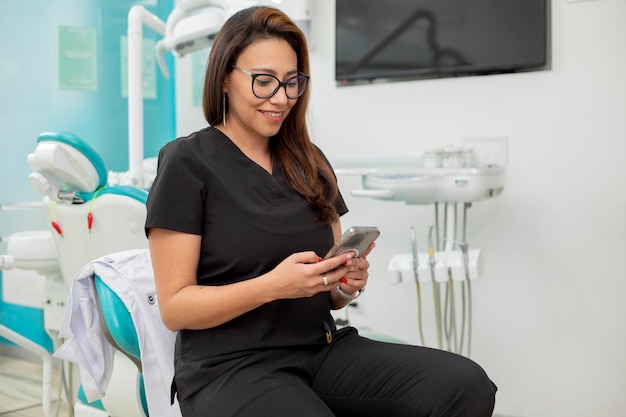 彼女のオフィスに座って携帯電話をチェックしている若いラテン系歯医者