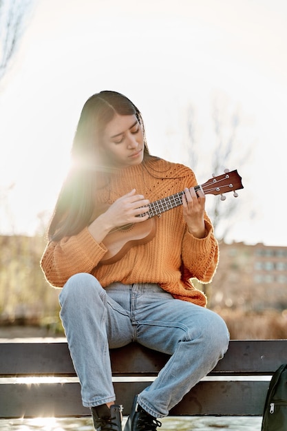 도시 공원에서 우쿨렐레를 연주하는 젊은 라티나 여성은 악기로 연습하는 벤치에 앉아 있다