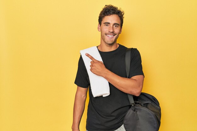 Young Latino athlete with backpack on yellow smiling and pointing aside showing something at blank space