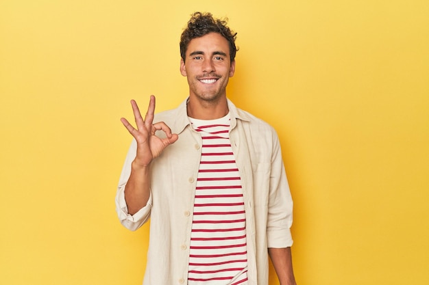 Young latino man posing on yellow background cheerful and confident showing ok gesture