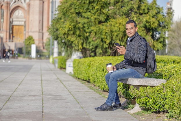 Young Latino man sitting on a bench in a public park using his mobile phone