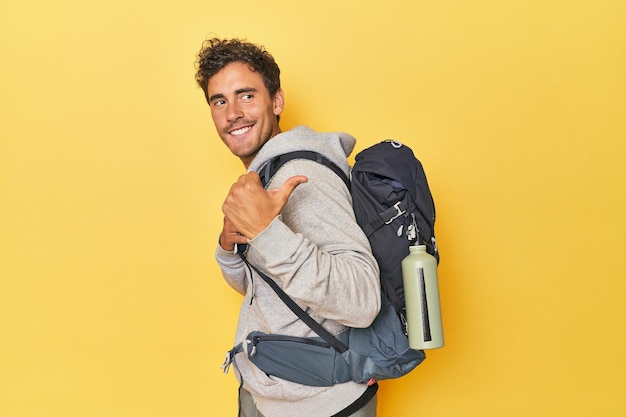 Young Latino with mountain backpack on yellow points with thumb finger away laughing and carefree