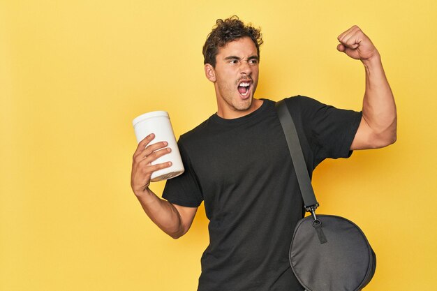 Young Latino with protein bottle on yellow raising fist after a victory winner concept