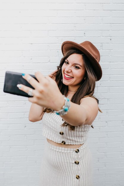 Young laughing attractive woman in hat taking photo on black mobile phone on white background