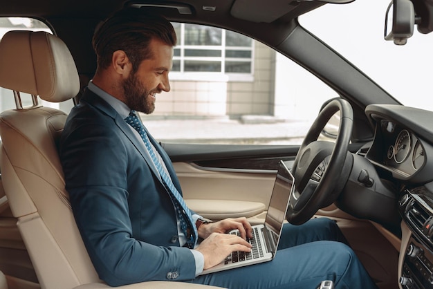 Young laughing businessman in suit using computer and internet in the automobile