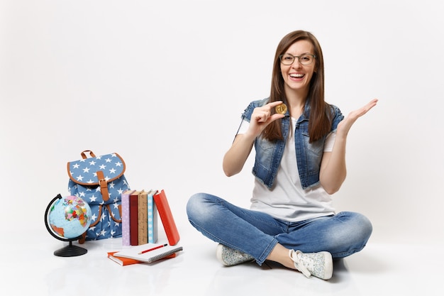 La giovane studentessa felice che ride con gli occhiali tiene in mano bitcoin che si allargano le mani siedono vicino al globo, zaino, libri di scuola isolati