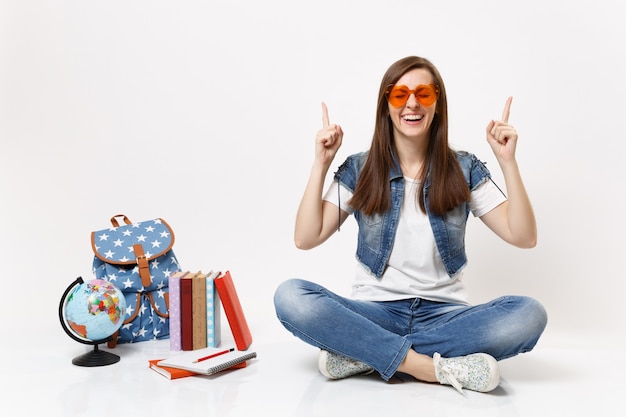 Young laughing happy woman student with closed eyes in red heart glasses pointing index fingers up near globe backpack books isolated