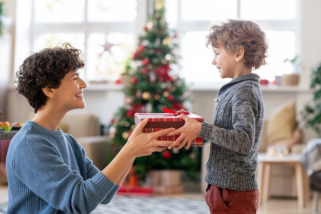 Photo young laughing mother passing christmas gift to her adorable little son while congratulating him on winter holiday in home environment
