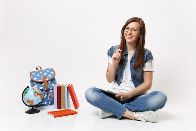 Foto giovane studentessa graziosa che ride con gli occhiali che tiene matita e taccuino seduto vicino al globo, zaino, libri scolastici isolati