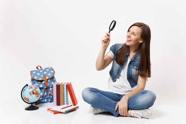 Young laughing pretty woman student holding looking on magnifying glass sitting near globe, backpack, school books isolated