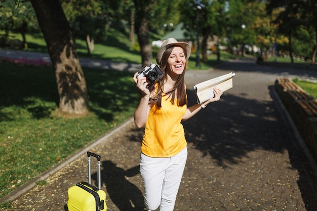 Фото Молодая смеющаяся туристическая женщина путешественника в шляпе с чемоданом, карта города, держащая ретро винтаж фотоаппарат в открытом городе. девушка едет за границу, чтобы поехать на выходные. туризм путешествие образ жизни.