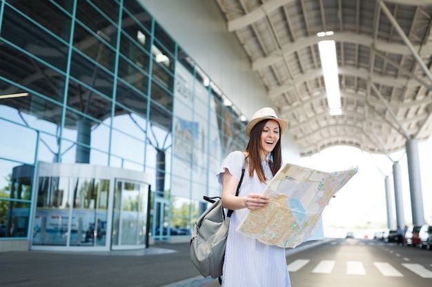 Young laughing traveler tourist woman with backpack holding paper map at international airport