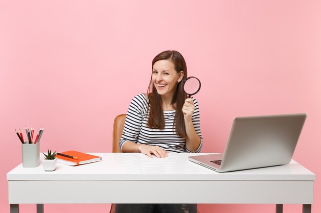 Young laughing woman in casual clothes holding magnifying glass sit working on project at white desk with pc laptop isolated on pastel pink background. Achievement business career concept. Copy space.