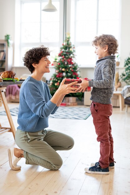 魔法の冬の休日に彼を祝福しながら彼女の愛らしい幼い息子にクリスマスプレゼントとボックスを渡す若い笑う女性