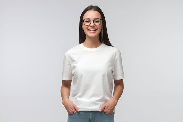 Young laughing woman standing with hands in pockets wearing blank white tshirt with copy space isolated on gray background