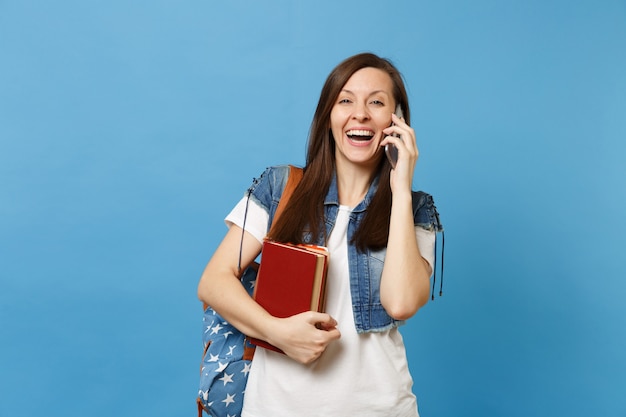 Молодой смеющийся студент женщины с рюкзаком держит школьные учебники, разговаривает по мобильному телефону, проводя приятный разговор, изолированный на синем фоне. Образование в концепции колледжа университета средней школы.