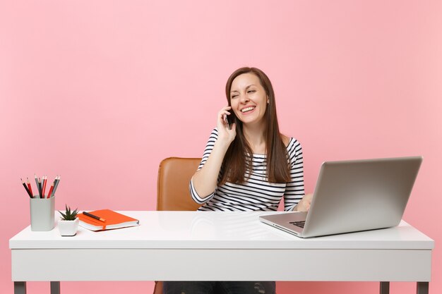 Young laughing woman talking on mobile phone, conducting pleasant conversation sit, work on project with pc laptop isolated on pastel pink background. Achievement business career concept. Copy space.