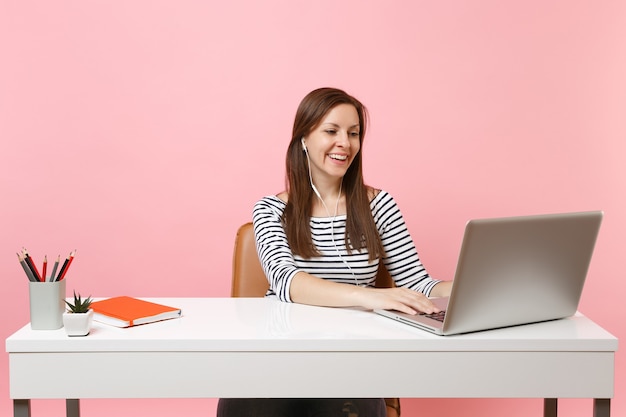 Young laughing woman with earphones listening music sitting and working at white desk with contemporary pc laptop 