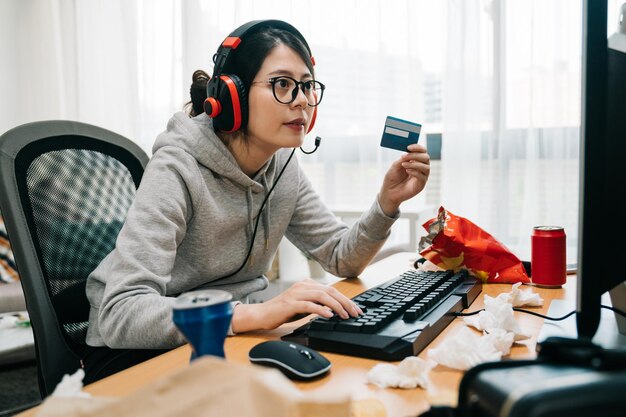 여름방학 동안 집에 있는 게으른 젊은 여대생 괴상한 것은 온라인 전자 상거래에서 게임 포인트를 구입하는 신용 카드를 들고 학교에서 쉬었습니다. 헤드폰을 끼고 컴퓨터 인터넷을 하는 아시아 여성 괴짜.