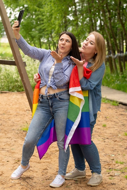 Young lesbian LGBT couple smiles while holding rainbow Pride flag LGBTQ selfie