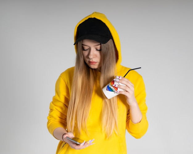Young LGBTQ sad woman wearing yellow hoodie jacket holding a cup with rainbow heart, looks into a smartphone, on gray background