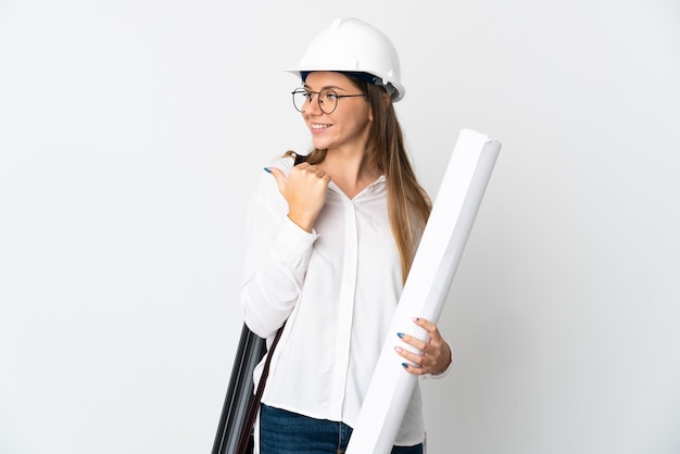 Young Lithuanian architect woman with helmet and holding blueprints isolated on white background pointing to the side to present a product