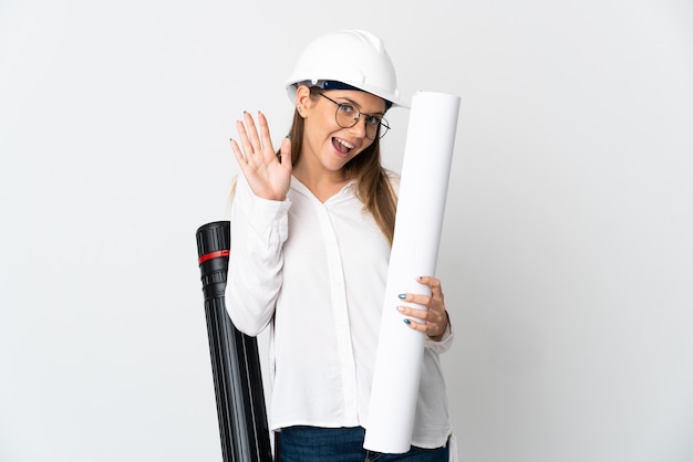 Young Lithuanian architect woman with helmet and holding blueprints isolated on white wall saluting with hand with happy expression