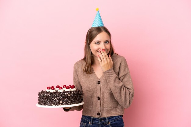 Giovane donna lituana che tiene la torta di compleanno isolata su sfondo rosa felice e sorridente che copre la bocca con la mano