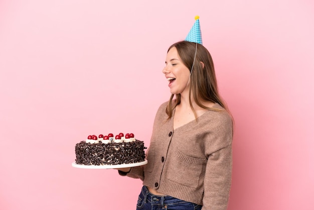 Молодая литовская женщина держит торт ко дню рождения на розовом фоне и смеется в боковом положении