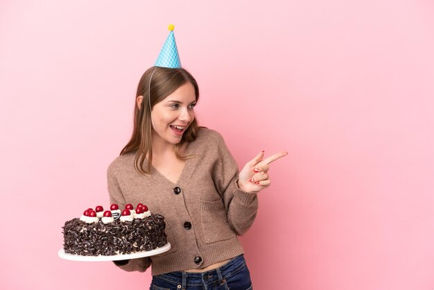 Молодая литовская женщина держит торт ко дню рождения на розовом фоне, указывая пальцем в сторону и представляя продукт