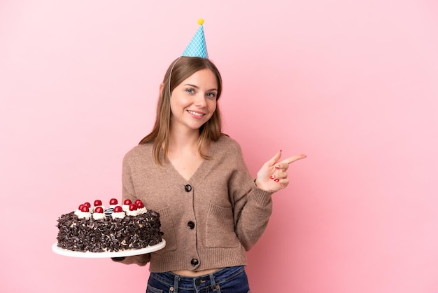 Young Lithuanian woman holding birthday cake isolated on pink background pointing finger to the side