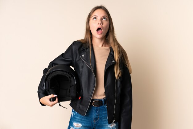 Young Lithuanian woman holding a motorcycle helmet isolated on beige wall looking up and with surprised expression