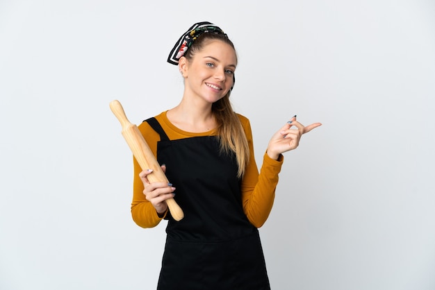 Young Lithuanian woman holding a rolling pin isolated on white wall pointing finger to the side