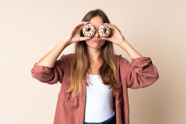 Young Lithuanian woman isolated on beige wall holding donuts in eyes with sad expression