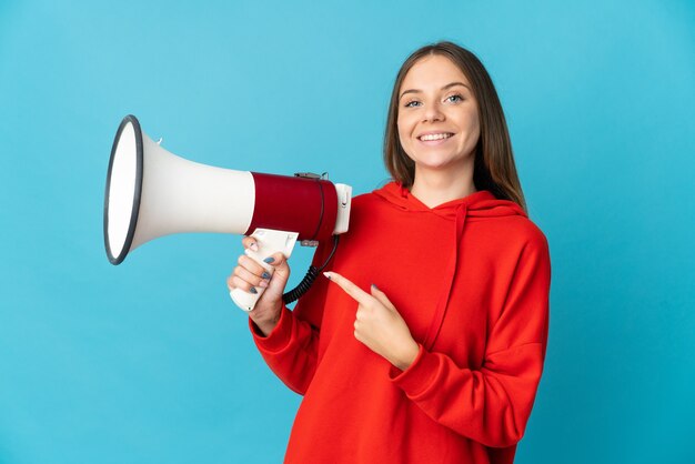 Young Lithuanian woman isolated on blue wall holding a megaphone and pointing side