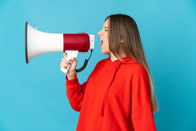 Young Lithuanian woman isolated on blue wall shouting through a megaphone to announce something in lateral position