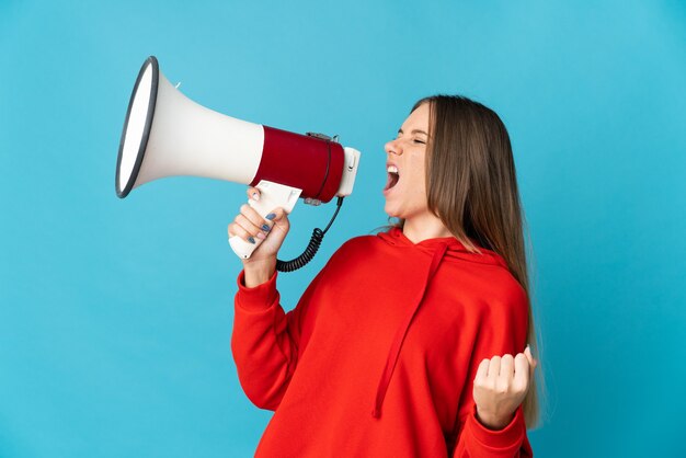 Young Lithuanian woman isolated on blue wall shouting through a megaphone to announce something in lateral position