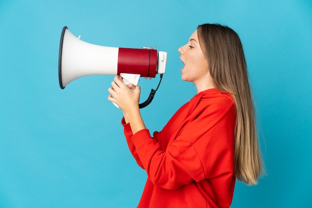 Young Lithuanian woman isolated on blue wall shouting through a megaphone to announce something in lateral position