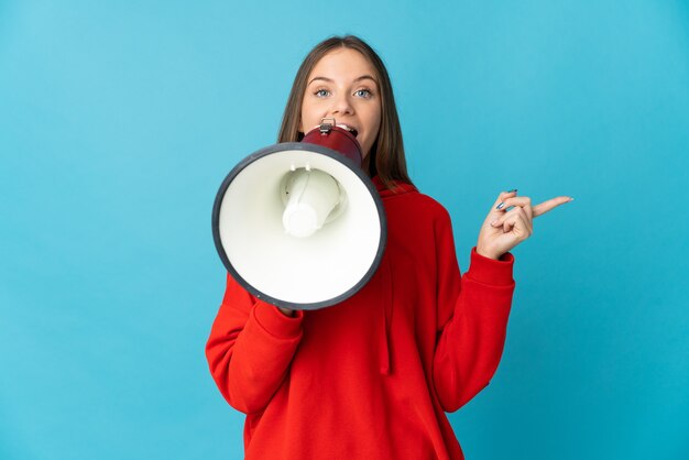 Young Lithuanian woman isolated on blue wall shouting through a megaphone and pointing side