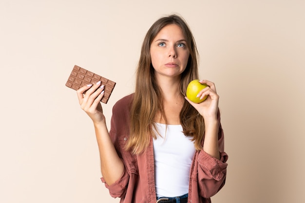 Фото Молодая литовская женщина изолирована на бежевом фоне, сомневаясь, принимая шоколадную таблетку в одной руке и яблоко в другой
