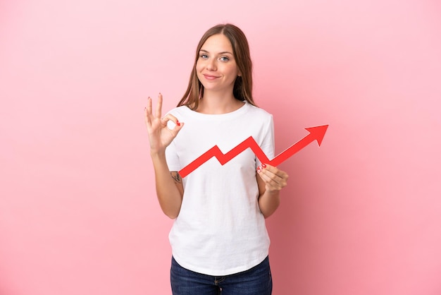 Young Lithuanian woman isolated on pink background holding a catching a rising arrow and doing OK sign